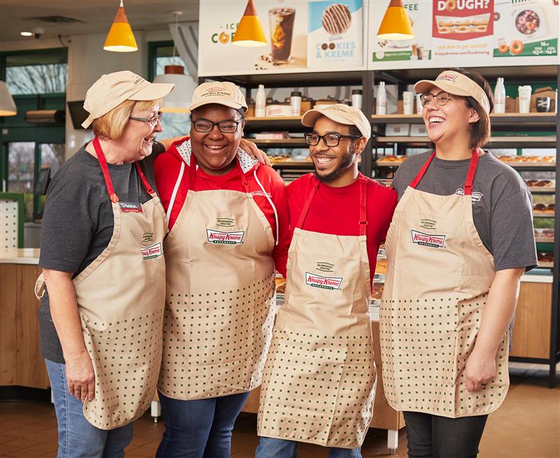 Four (4) Krispy Kreme employees smiling inside doughnut shop